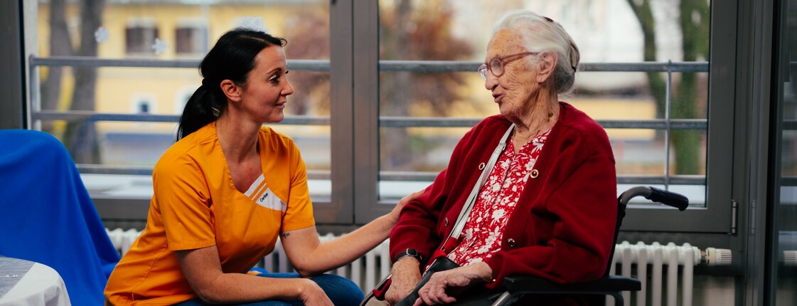 Links eine junge Frau in orangem Shirt sitzt und hält den Arm einer alten Dame, die im Rollstuhl sitzt.